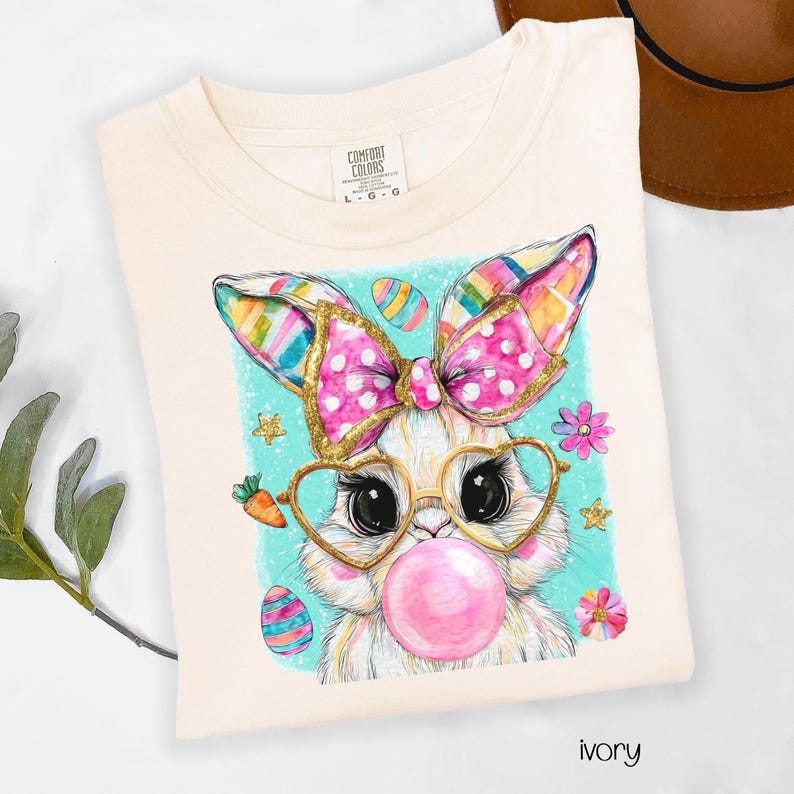 Peut inclure: T-shirt ivoire avec un motif de lapin de P&acirc;ques fantaisiste. Le lapin porte des lunettes en forme de c&oelig;ur et un n&oelig;ud rose, soufflant une bulle de chewing-gum. Des &oelig;ufs de P&acirc;ques et des fleurs color&eacute;s entourent le lapin sur un fond turquoise.