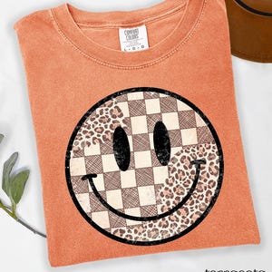May include: Terracotta-colored t-shirt with a large smiley face graphic. The smiley face is divided into sections of checkered pattern and leopard print. The shirt has a crew neck and short sleeves. The text "terracotta" is at the bottom.