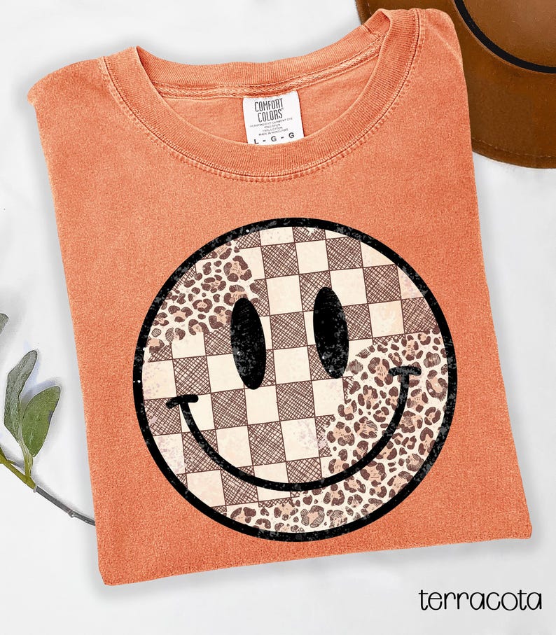 May include: Terracotta-colored t-shirt with a large smiley face graphic. The smiley face is divided into sections of checkered pattern and leopard print. The shirt has a crew neck and short sleeves. The text "terracotta" is at the bottom.