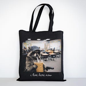 May include: Black tote bag featuring a raccoon wearing sunglasses and a denim cap, holding a drink with a straw. The raccoon is seated in a classroom. The text "i live here now" is written in cursive at the bottom of the image.
