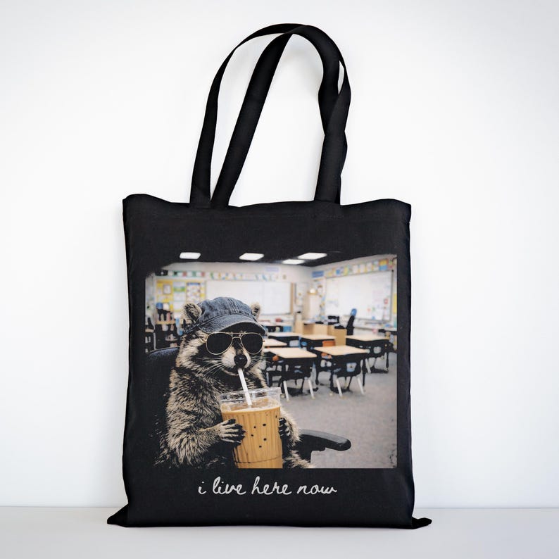 May include: Black tote bag featuring a raccoon wearing sunglasses and a denim cap, holding a drink with a straw. The raccoon is seated in a classroom. The text "i live here now" is written in cursive at the bottom of the image.