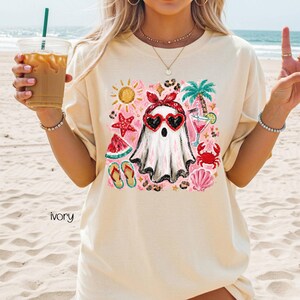 May include: Ivory-colored t-shirt featuring a whimsical ghost design. The ghost wears heart-shaped sunglasses and a red headband, surrounded by summer-themed illustrations like a watermelon slice, flip-flops, and a cocktail. The word "ivory" is printed at the bottom.