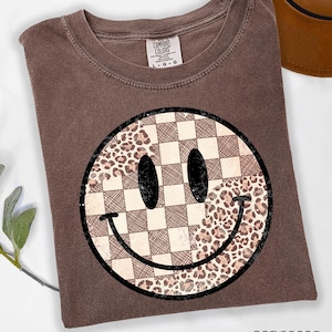 May include: Espresso-colored t-shirt featuring a large smiley face design. The smiley face is divided into sections with a checkered pattern and a leopard print pattern. The shirt has a crew neck and short sleeves.