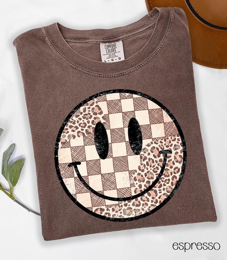 May include: Espresso-colored t-shirt featuring a large smiley face design. The smiley face is divided into sections with a checkered pattern and a leopard print pattern. The shirt has a crew neck and short sleeves.