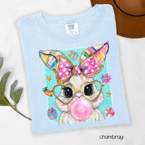 Peut inclure: T-shirt bleu clair Comfort Colors avec un lapin de dessin anim&eacute; portant des lunettes en forme de c&oelig;ur et soufflant une bulle. Le lapin a un n&oelig;ud rose &agrave; pois blancs et des oreilles color&eacute;es. Th&egrave;me de P&acirc;ques avec des &oelig;ufs, des carottes et des fleurs.
