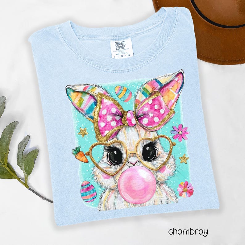 Peut inclure: T-shirt bleu clair Comfort Colors avec un lapin de dessin anim&eacute; portant des lunettes en forme de c&oelig;ur et soufflant une bulle. Le lapin a un n&oelig;ud rose &agrave; pois blancs et des oreilles color&eacute;es. Th&egrave;me de P&acirc;ques avec des &oelig;ufs, des carottes et des fleurs.