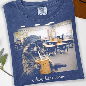 May include: Denim blue t-shirt featuring a raccoon wearing sunglasses and a cap, holding a drink with a straw. The image inside the frame shows a classroom. Text reads "i live here now".