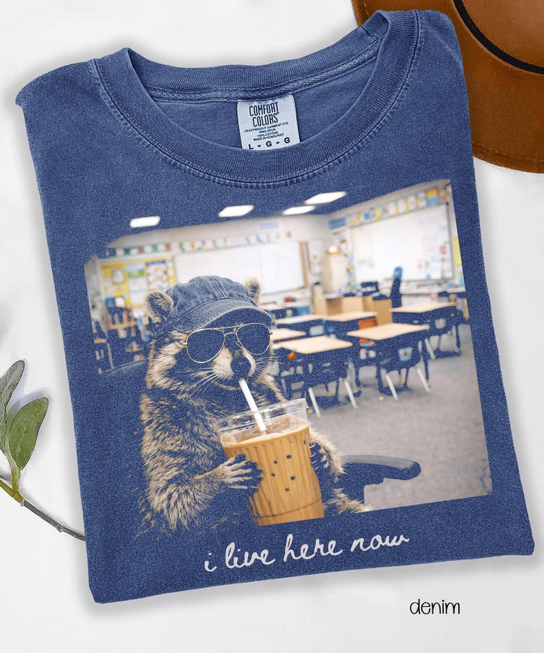 May include: Denim blue t-shirt featuring a raccoon wearing sunglasses and a cap, holding a drink with a straw. The image inside the frame shows a classroom. Text reads "i live here now".