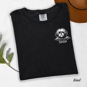 Puede incluir: Camiseta negra con un gr&aacute;fico blanco de un perro Shih Tzu y el nombre "Bella" impreso debajo. La camiseta es de la marca Comfort Colors y est&aacute; colocada sobre una superficie blanca.