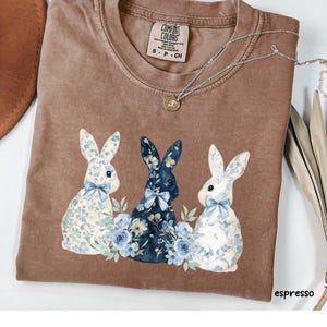 Puede incluir: Una camiseta marrón con un gráfico de tres conejos florales. Los conejos llevan lazos florales azules y blancos. La camiseta tiene el texto "COMFORT COLORS" y "S - P - CH" en la etiqueta.