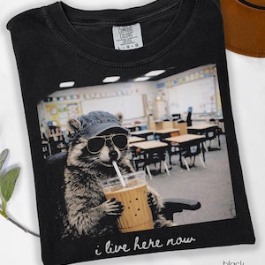 May include: Black t-shirt featuring a raccoon wearing sunglasses and a hat, sipping from a beverage cup. The image on the shirt shows a classroom setting with the text "i live here now". The shirt is labeled "Comfort Colors".