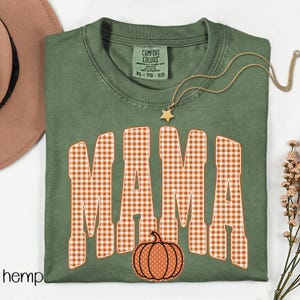 Pode incluir: Uma t-shirt verde-oliva com a palavra MAMA num padr&atilde;o xadrez branco e laranja. Uma pequena ab&oacute;bora laranja com pintas brancas est&aacute; posicionada abaixo do texto. A t-shirt tem uma etiqueta Comfort Colors e um colar com pendente de estrela.
