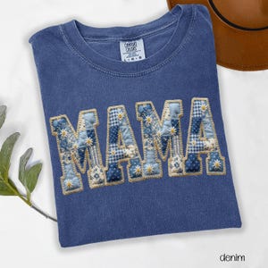 May include: A blue denim t-shirt with the word "MAMA" appliqued across the front. The letters are made of patchwork fabric in various shades of blue and white, with small daisy accents. The shirt has a crew neck and short sleeves.