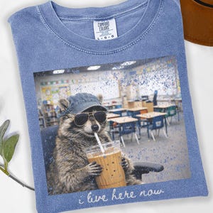 May include: Blue jean Comfort Colors t-shirt featuring a raccoon wearing sunglasses and a cap, holding a coffee drink with a straw. The graphic includes the text "i live here now". The shirt is a light blue color.