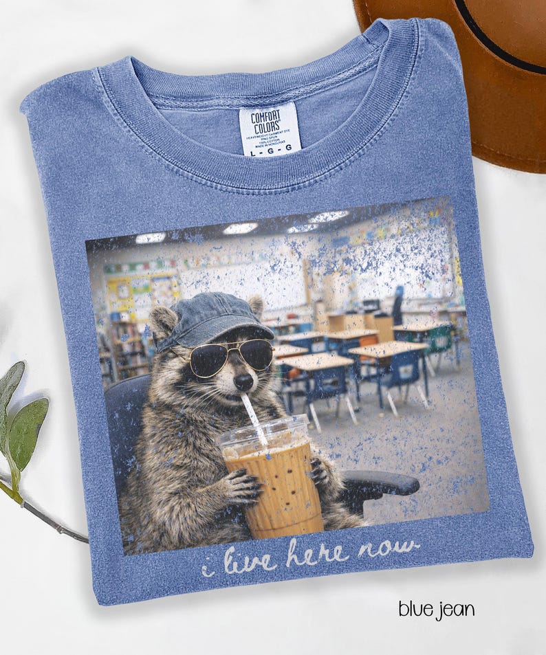 May include: Blue jean Comfort Colors t-shirt featuring a raccoon wearing sunglasses and a cap, holding a coffee drink with a straw. The graphic includes the text "i live here now". The shirt is a light blue color.