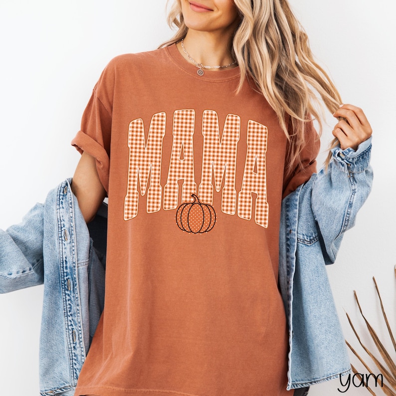 Pode incluir: Uma t-shirt de manga curta cor de ferrugem com a palavra MAMA num padr&atilde;o de xadrez vichy branco e laranja. Abaixo do texto encontra-se um gr&aacute;fico de uma ab&oacute;bora com uma textura de pontos. A t-shirt &eacute; usada sob um casaco de ganga azul claro.