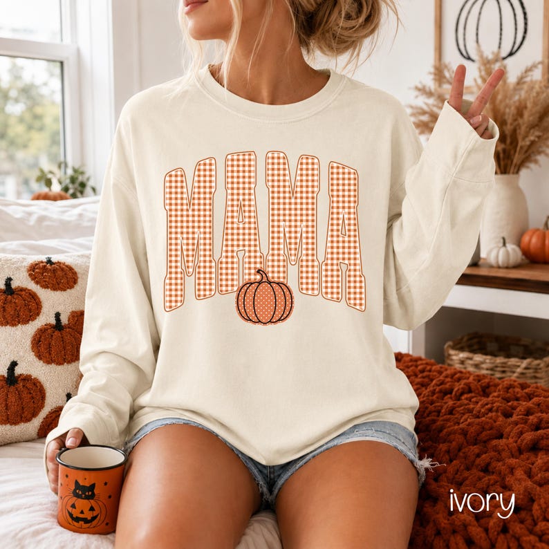 Pode incluir: Uma camisola de manga comprida cor de marfim com a palavra MAMA num padr&atilde;o xadrez vichy laranja e branco. A letra A central &eacute; substitu&iacute;da por um gr&aacute;fico de ab&oacute;bora laranja com bolinhas brancas. A camisola tem um decote redondo.