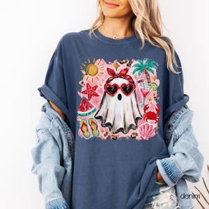 May include: A navy blue t-shirt featuring a graphic of a ghost wearing heart-shaped sunglasses and a red bandana, surrounded by summer-themed illustrations. The design includes a sun, palm tree, watermelon, and cocktail.