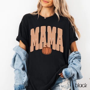 Pode incluir: Uma t-shirt preta de manga curta com a palavra MAMA numa fonte grande de estilo universit&aacute;rio aos quadrados cor de laranja e brancos. Um gr&aacute;fico de ab&oacute;bora cor de laranja com pintas brancas est&aacute; posicionado abaixo do texto na frente.