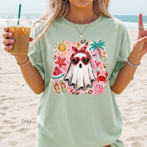 May include: A light green t-shirt featuring a whimsical ghost design. The ghost wears heart-shaped sunglasses and a red bandana, surrounded by summer-themed illustrations like a sun, palm tree, and watermelon. The word "bay" is printed on the shirt.
