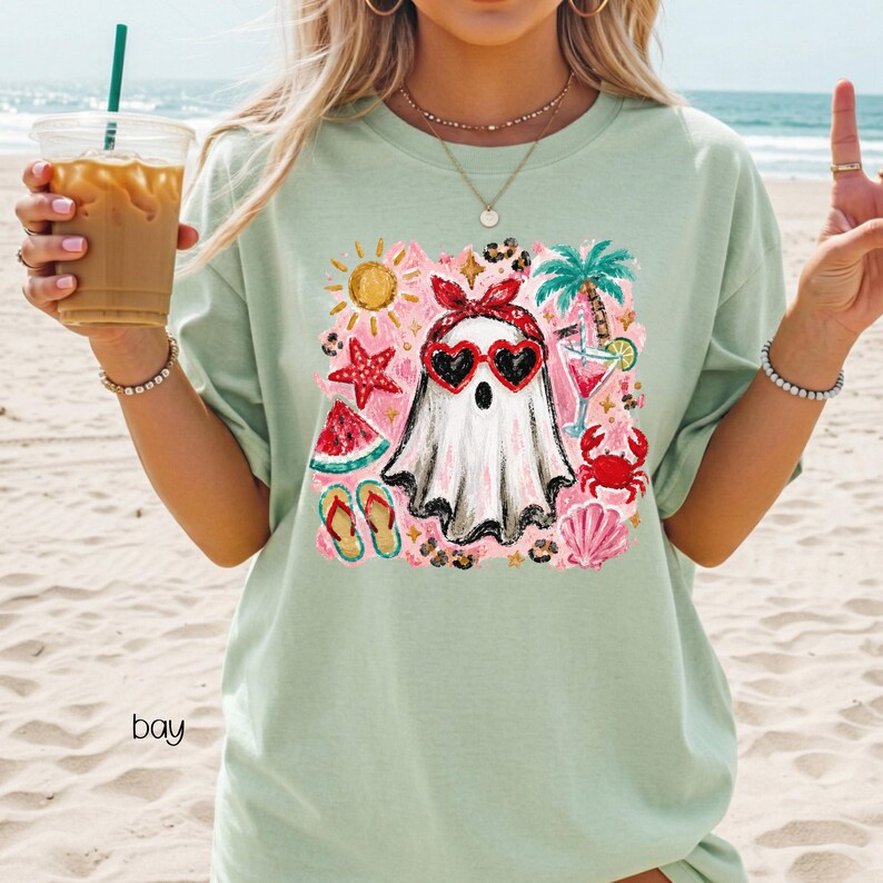 May include: A light green t-shirt featuring a whimsical ghost design. The ghost wears heart-shaped sunglasses and a red bandana, surrounded by summer-themed illustrations like a sun, palm tree, and watermelon. The word "bay" is printed on the shirt.