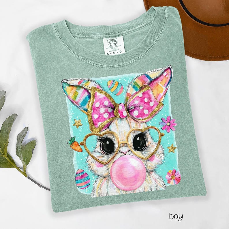 Peut inclure: T-shirt vert clair Comfort Colors avec un motif de lapin de dessin anim&eacute;. Le lapin porte des lunettes en forme de c&oelig;ur et fait une bulle de chewing-gum. Il a un n&oelig;ud rose &agrave; pois et des oreilles color&eacute;es. Le mot "bay" est imprim&eacute; en bas.