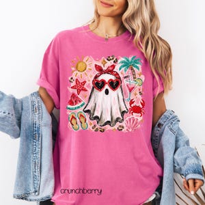 May include: Pink t-shirt featuring a graphic of a ghost wearing heart-shaped sunglasses and a red bandana, surrounded by summer-themed illustrations. The shirt has rolled-up sleeves and the text "crunchberry" at the bottom.