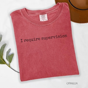 May include: Crimson-colored t-shirt with the text "I require supervision" in black. The shirt is a Comfort Colors brand and is folded neatly. A brown hat is partially visible in the background.
