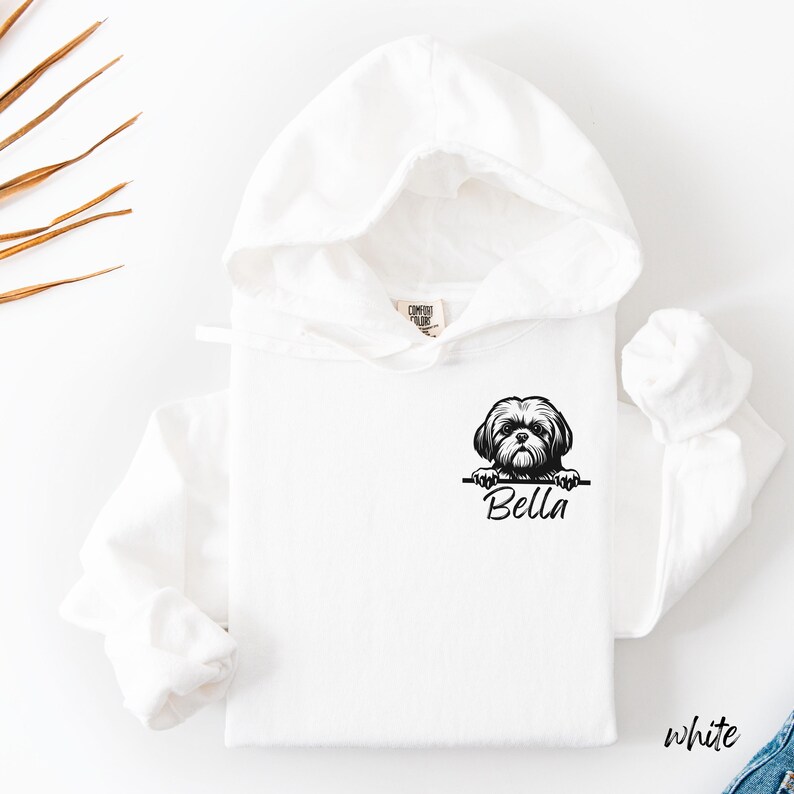 Puede incluir: Sudadera con capucha blanca con un gr&aacute;fico de perro Shih Tzu en blanco y negro y el nombre "Bella" impreso debajo. La sudadera tiene mangas largas y una capucha con cord&oacute;n. La palabra "white" est&aacute; impresa en la esquina inferior derecha.