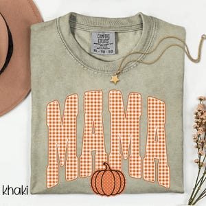 Pode incluir: Uma t-shirt de gola redonda na cor c&aacute;qui com a palavra MAMA em letras grandes de bloco com padr&atilde;o xadrez laranja e branco. Abaixo do texto, h&aacute; um pequeno gr&aacute;fico de uma ab&oacute;bora laranja com bolinhas brancas. A t-shirt tem uma etiqueta Comfort Colors.