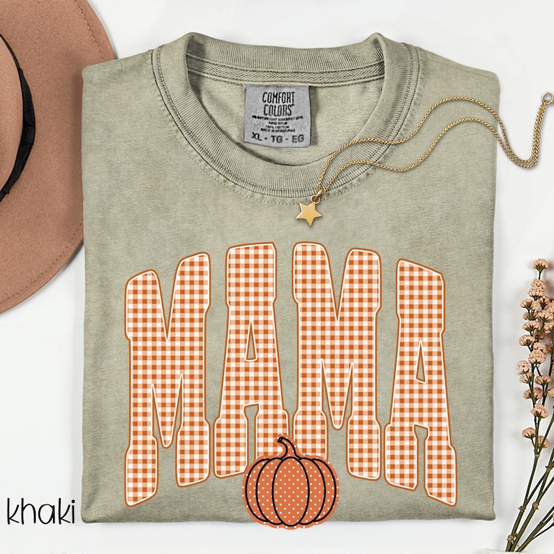 Pode incluir: Uma t-shirt de gola redonda na cor c&aacute;qui com a palavra MAMA em letras grandes de bloco com padr&atilde;o xadrez laranja e branco. Abaixo do texto, h&aacute; um pequeno gr&aacute;fico de uma ab&oacute;bora laranja com bolinhas brancas. A t-shirt tem uma etiqueta Comfort Colors.