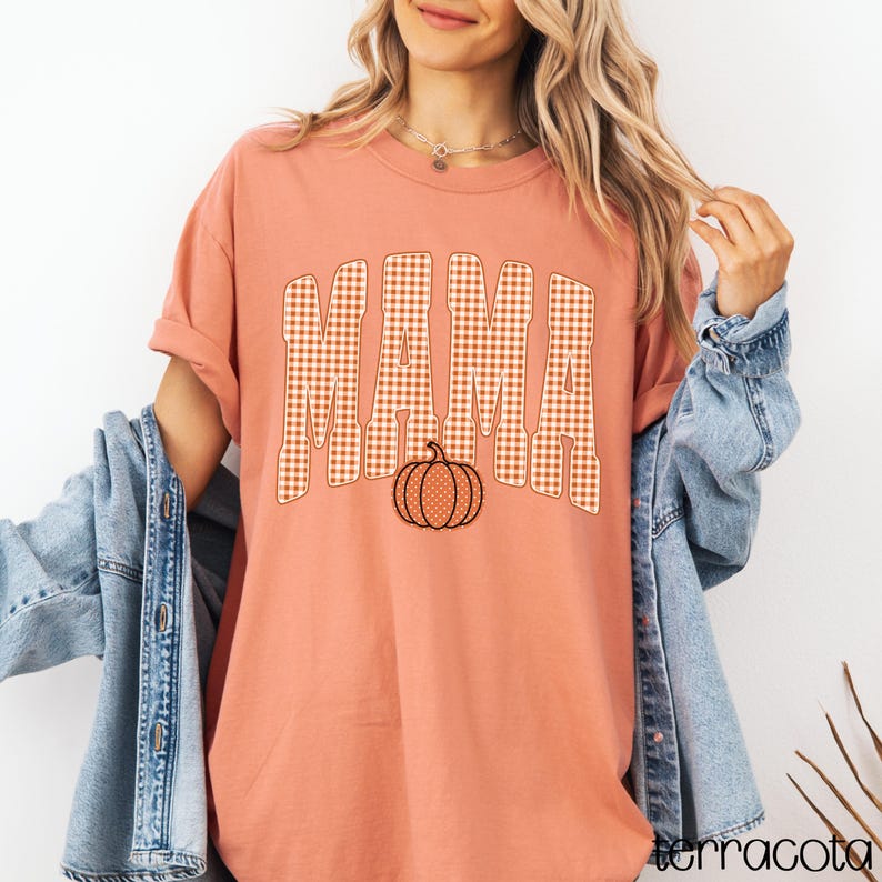Pode incluir: Uma t-shirt de cor terracota com a palavra MAMA num padr&atilde;o xadrez vichy laranja e branco. Abaixo do texto encontra-se um gr&aacute;fico de uma ab&oacute;bora com um padr&atilde;o de bolinhas brancas. A t-shirt &eacute; usada com um casaco de ganga azul claro.