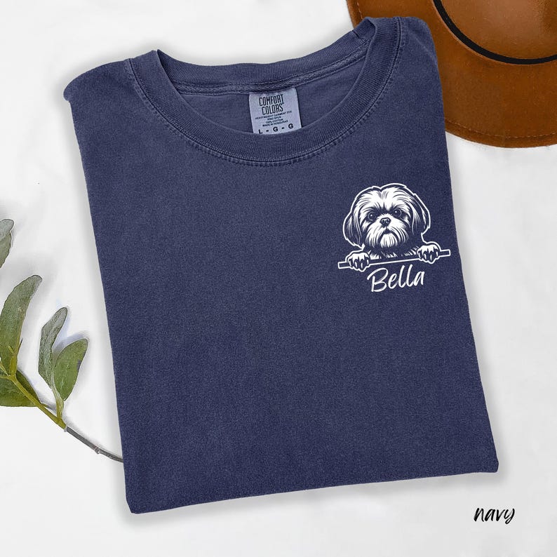 Puede incluir: Camiseta azul marino con un gr&aacute;fico blanco de un perro Shih Tzu y el nombre "Bella". La camiseta tiene cuello redondo y est&aacute; hecha de una tela suave. La etiqueta de la marca Comfort Colors es visible.