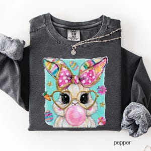 Peut inclure: Sweat-shirt gris fonc&eacute; avec un motif de lapin de dessin anim&eacute;. Le lapin porte des lunettes et fait une bulle de chewing-gum. Le motif comprend un n&oelig;ud rose, des &oelig;ufs de P&acirc;ques et des fleurs. Un collier argent&eacute; est drap&eacute; sur le chemisier.