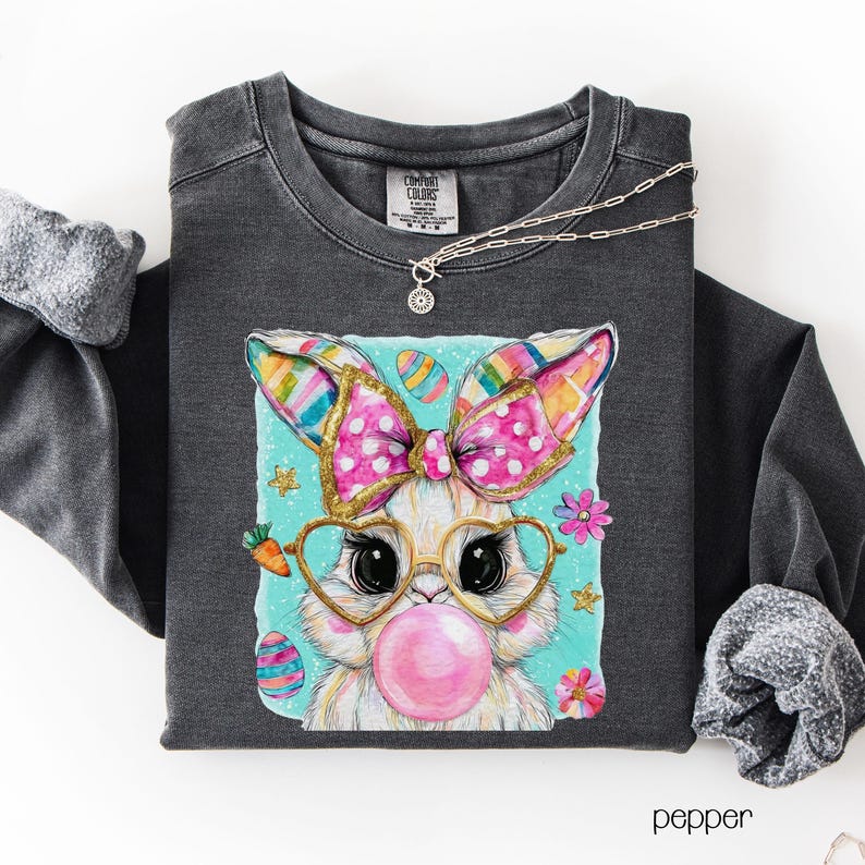 Peut inclure: Sweat-shirt gris fonc&eacute; avec un motif de lapin de dessin anim&eacute;. Le lapin porte des lunettes et fait une bulle de chewing-gum. Le motif comprend un n&oelig;ud rose, des &oelig;ufs de P&acirc;ques et des fleurs. Un collier argent&eacute; est drap&eacute; sur le chemisier.
