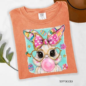 Peut inclure: T-shirt terracotta avec un motif de lapin de dessin anim&eacute; portant des lunettes en forme de c&oelig;ur et une bulle rose. Le motif comprend des &oelig;ufs de P&acirc;ques et des fleurs. Le t-shirt porte l'&eacute;tiquette "Comfort Colors".