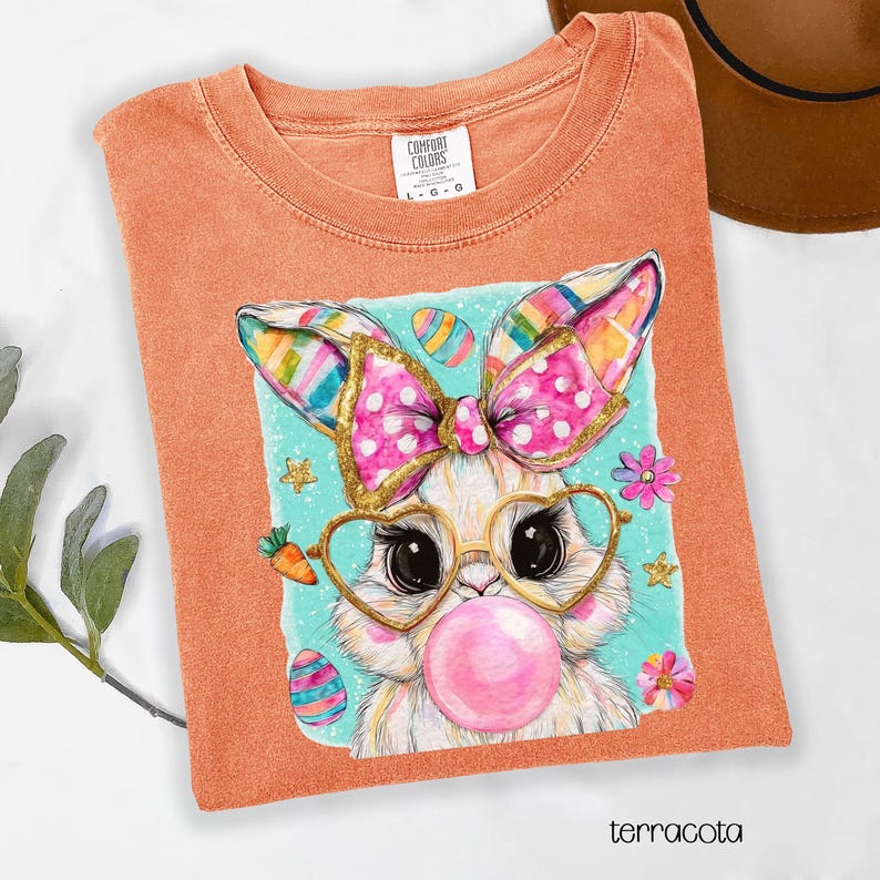 Peut inclure: T-shirt terracotta avec un motif de lapin de dessin anim&eacute; portant des lunettes en forme de c&oelig;ur et une bulle rose. Le motif comprend des &oelig;ufs de P&acirc;ques et des fleurs. Le t-shirt porte l'&eacute;tiquette "Comfort Colors".