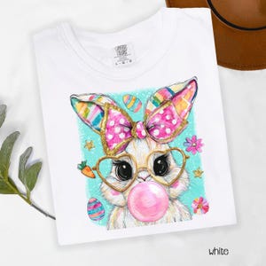 Peut inclure: T-shirt blanc avec un motif de lapin de dessin anim&eacute;. Le lapin porte des lunettes en forme de c&oelig;ur et fait une bulle de chewing-gum. Il a un n&oelig;ud rose avec des paillettes dor&eacute;es et des oreilles color&eacute;es. Des &oelig;ufs de P&acirc;ques et des fleurs sont sur un fond turquoise.