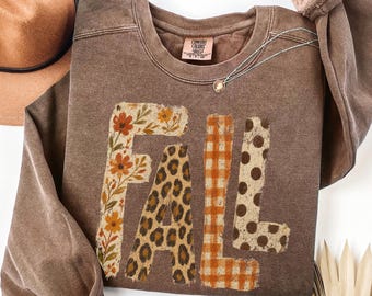 May include: A brown long-sleeve crewneck sweatshirt featuring the word FALL in large block letters. Each letter has a different pattern: the F is floral, the A is leopard print, the L is orange gingham, and the L is brown polka dots.