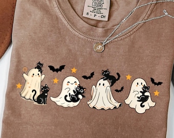 May include: An espresso-colored t-shirt featuring a graphic print of four white ghosts interacting with black cats, bats, and yellow stars. The shirt has a Comfort Colors brand label at the neck and a silver-toned necklace resting on the fabric.