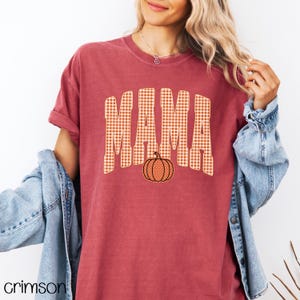 Pode incluir: Uma t-shirt de manga curta vermelho-escura com a palavra MAMA num padr&atilde;o de xadrez vichy bege e branco. Abaixo do texto encontra-se um pequeno gr&aacute;fico de uma ab&oacute;bora laranja com textura pontilhada. Usada com um casaco de ganga azul claro.