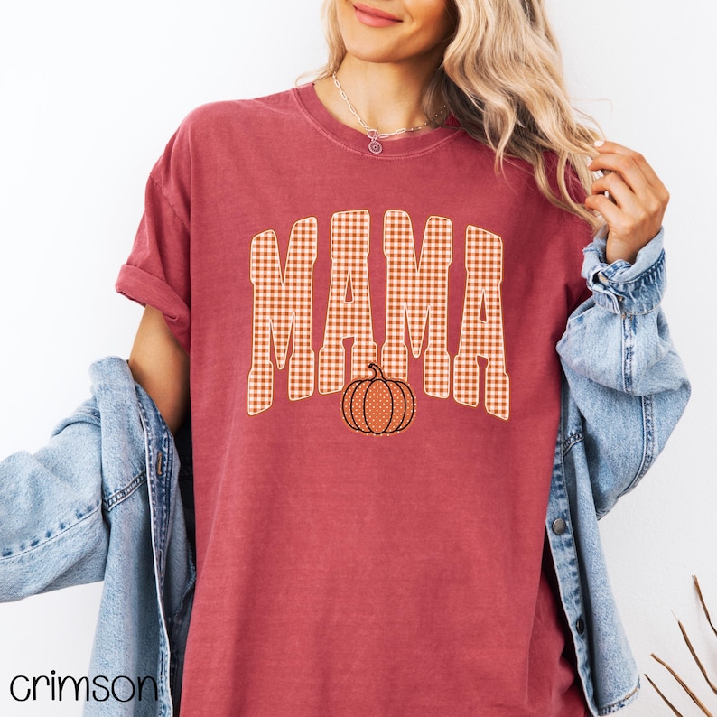 Pode incluir: Uma t-shirt de manga curta vermelho-escura com a palavra MAMA num padr&atilde;o de xadrez vichy bege e branco. Abaixo do texto encontra-se um pequeno gr&aacute;fico de uma ab&oacute;bora laranja com textura pontilhada. Usada com um casaco de ganga azul claro.