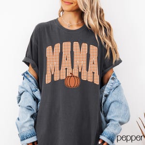 Pode incluir: Uma t-shirt de manga curta cinzento-carv&atilde;o com a palavra MAMA num padr&atilde;o grande de xadrez vichy laranja e branco. Um gr&aacute;fico de ab&oacute;bora laranja com bolinhas brancas est&aacute; posicionado diretamente abaixo do texto na frente da t-shirt.