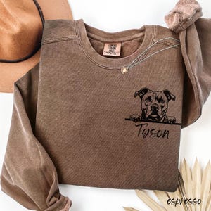May include: Espresso-colored sweatshirt featuring a black graphic of a dog with the name "Tyson" below. The sweatshirt has a crew neck and long sleeves. A silver necklace with a small pendant is visible.