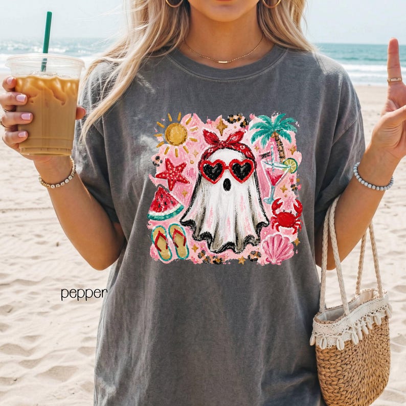 May include: Dark grey t-shirt featuring a graphic of a ghost wearing heart-shaped sunglasses and a red bow, surrounded by beach-themed illustrations. The word "pepper" is printed on the shirt. The design includes a sun, palm tree, and watermelon.