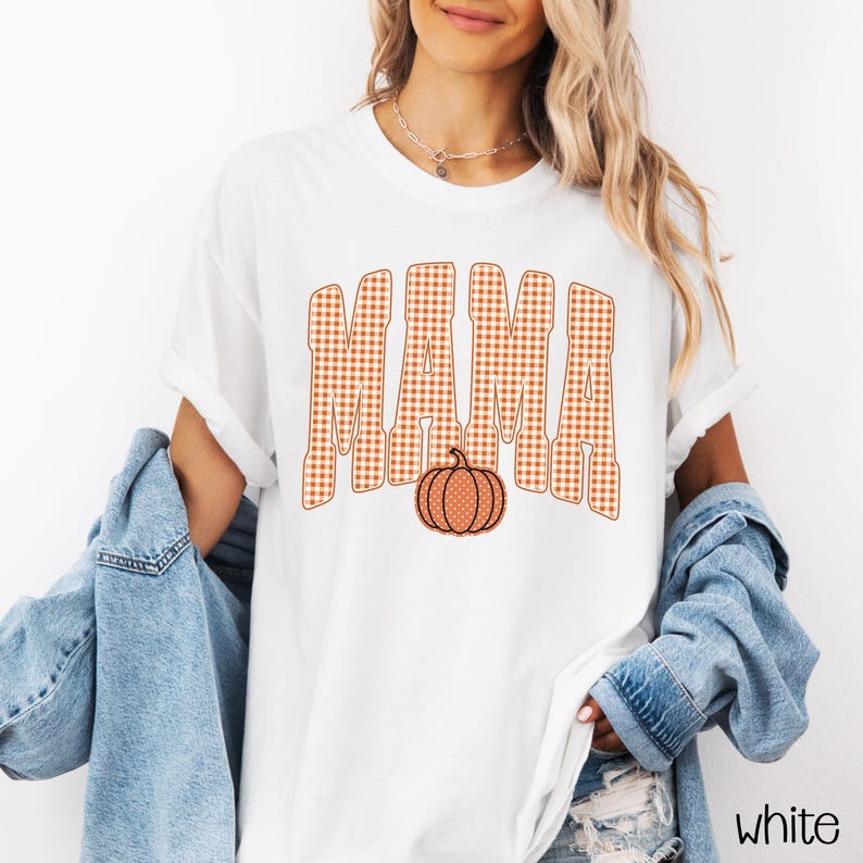 Pode incluir: Uma t-shirt branca de manga curta com a palavra MAMA numa fonte grande aos quadrados cor de laranja e brancos. Por baixo do texto, existe um gr&aacute;fico de uma ab&oacute;bora cor de laranja com um padr&atilde;o de bolinhas brancas. A t-shirt &eacute; usada com um blus&atilde;o de ganga azul.