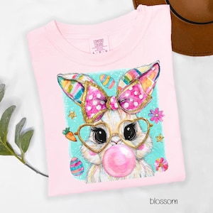 Peut inclure: T-shirt rose clair avec un motif de lapin de dessin anim&eacute;. Le lapin porte des lunettes en forme de c&oelig;ur et fait une bulle. Il a un n&oelig;ud rose avec des bordures dor&eacute;es et des oreilles color&eacute;es. Le mot "blossom" est imprim&eacute; en bas.