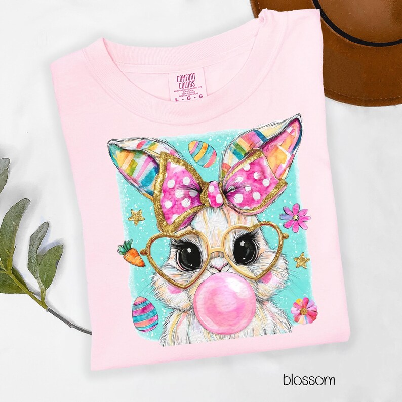 Peut inclure: T-shirt rose clair avec un motif de lapin de dessin anim&eacute;. Le lapin porte des lunettes en forme de c&oelig;ur et fait une bulle. Il a un n&oelig;ud rose avec des bordures dor&eacute;es et des oreilles color&eacute;es. Le mot "blossom" est imprim&eacute; en bas.