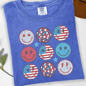 Pode incluir: Uma t-shirt azul jeans com etiqueta Comfort Colors apresentando uma estampa gráfica de nove designs circulares, incluindo rostos sorridentes, bolas de discoteca e padrões da bandeira americana, acentuados com pequenas estrelas em acabamento vintage.