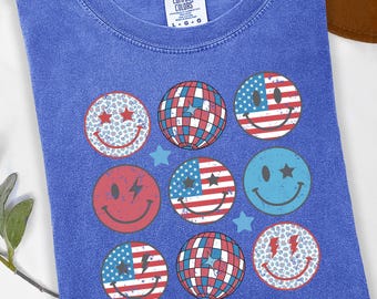 Peut inclure: Un t-shirt bleu jean avec une étiquette Comfort Colors présentant un imprimé graphique de neuf motifs circulaires incluant des visages souriants, des boules à facettes et des drapeaux américains, agrémenté de petites étoiles au fini vieilli.